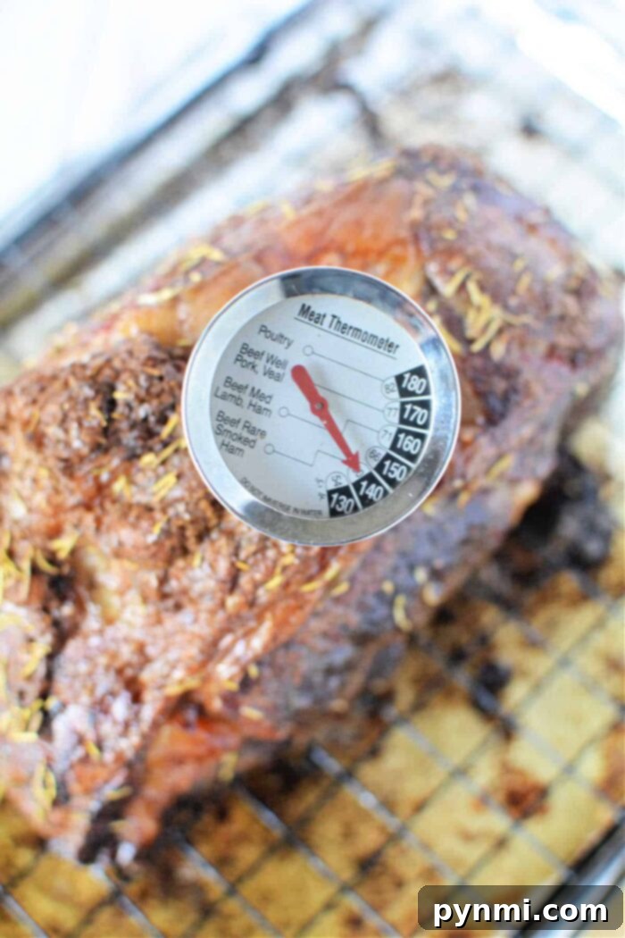 A meat thermometer is inserted into a cooked boneless rib roast, showing a temperature reading of medium-rare. The roast, seasoned with herbs, rests on a wire rack set on a baking sheet. The thermometer's dial displays various meat types and corresponding temperature ranges, emphasizing the importance of precision in achieving the perfect doneness.