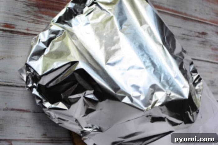 A piece of food, possibly involving a boneless rib roast recipe, is wrapped in crinkled aluminum foil on a wooden surface. The aluminum foil reflects light and covers the object completely. This depicts the crucial resting phase of the roast, where it continues to cook and juices redistribute for maximum tenderness.