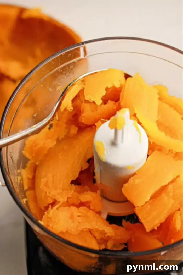 Crockpot Pumpkin Perfection 7 A food processor bowl is filled with chunks of cooked pumpkin. A metal spoon rests on the edge of the bowl. The pieces are bright orange and partially mashed, blending flavors beautifully. The background shows more sweet potato peels, indicating a festive preparation.