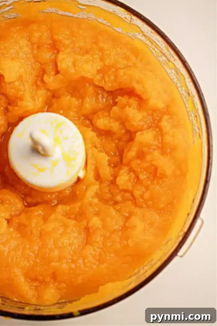 Crockpot Pumpkin Perfection 8 A food processor bowl filled with smooth, bright orange puree from pumpkin cooked in a slow cooker. The puree is evenly blended, and the white blade of the food processor is visible in the center. The sides of the bowl are coated with some of the mixture.