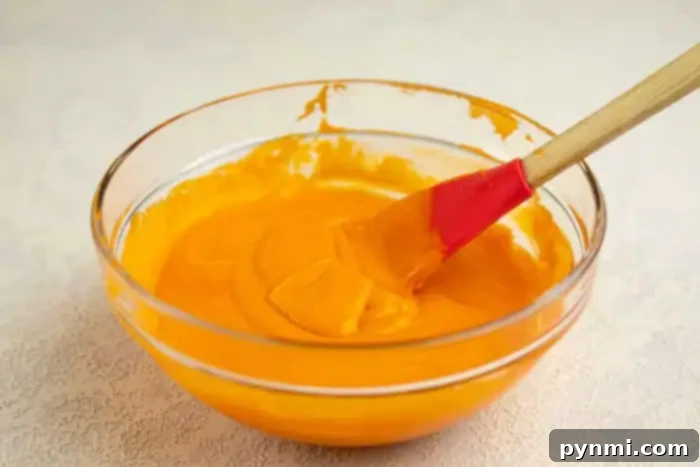 Surprise Pumpkin Loaf Cake 3 A clear glass bowl, carefully shaped and containing a smooth, orange mixture, is shown. A red and wooden spatula rests inside the bowl, partially covered with the mixture. The bowl is placed on a light-colored, textured surface.