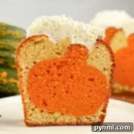 A single piece of cake on a white plate reveals an orange pumpkin shape inside the cake. Topped with a layer of white frosting, the cross-section showcases the charming design. In the blurred background, another piece of cake awaits its turn.