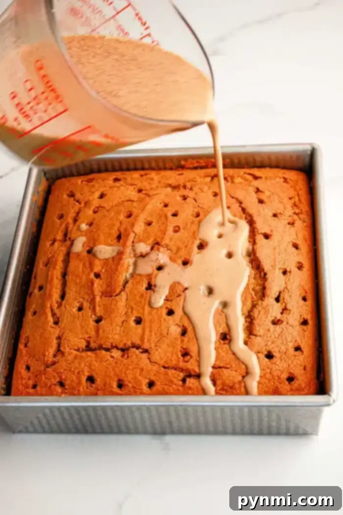 A square Pumpkin Poke Cake in a metal baking pan is being drizzled with a light brown liquid from a measuring cup. The cake has multiple holes poked into its surface to allow the liquid to seep in, ensuring every bite is moist and flavorful.