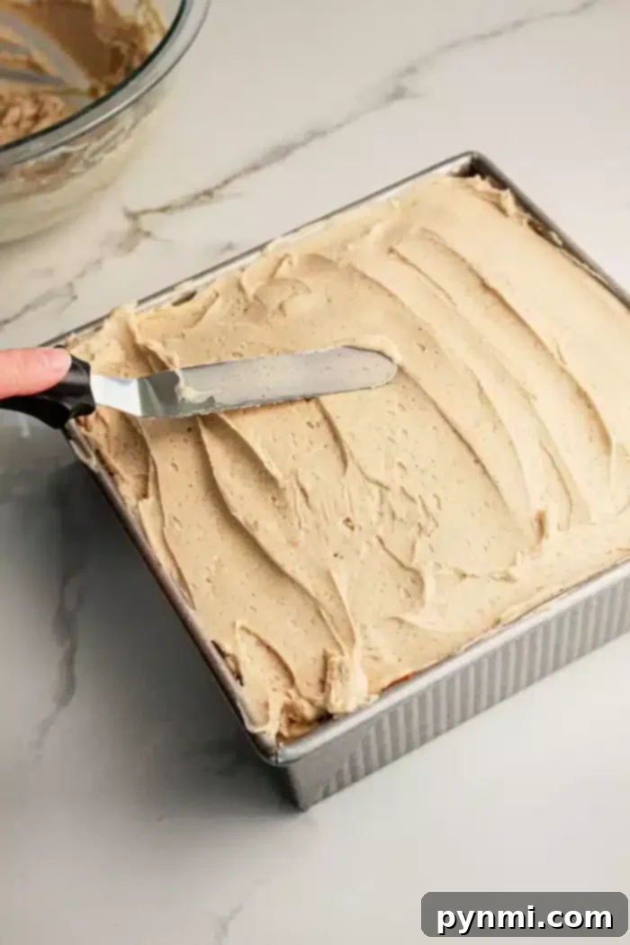 A hand holding a straight icing spatula is spreading beige frosting evenly over a square Pumpkin Poke Cake in a metal baking pan. The pan is sitting on a marble countertop, and a glass bowl with additional frosting is visible in the background.