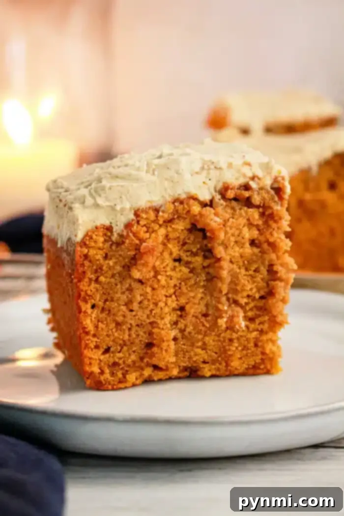 A close-up of a slice of Pumpkin Poke Cake with cream cheese frosting on a white plate. The cake has a golden-brown color and a moist texture, while the thick frosting covers the top of the slice. A blurred background shows another slice and a lit candle.