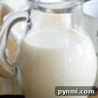 A clear glass pitcher filled with creamer sits gracefully on a wooden table, paired with a white and yellow cloth nearby, ready to transform into homemade coffee creamer for your morning brew.