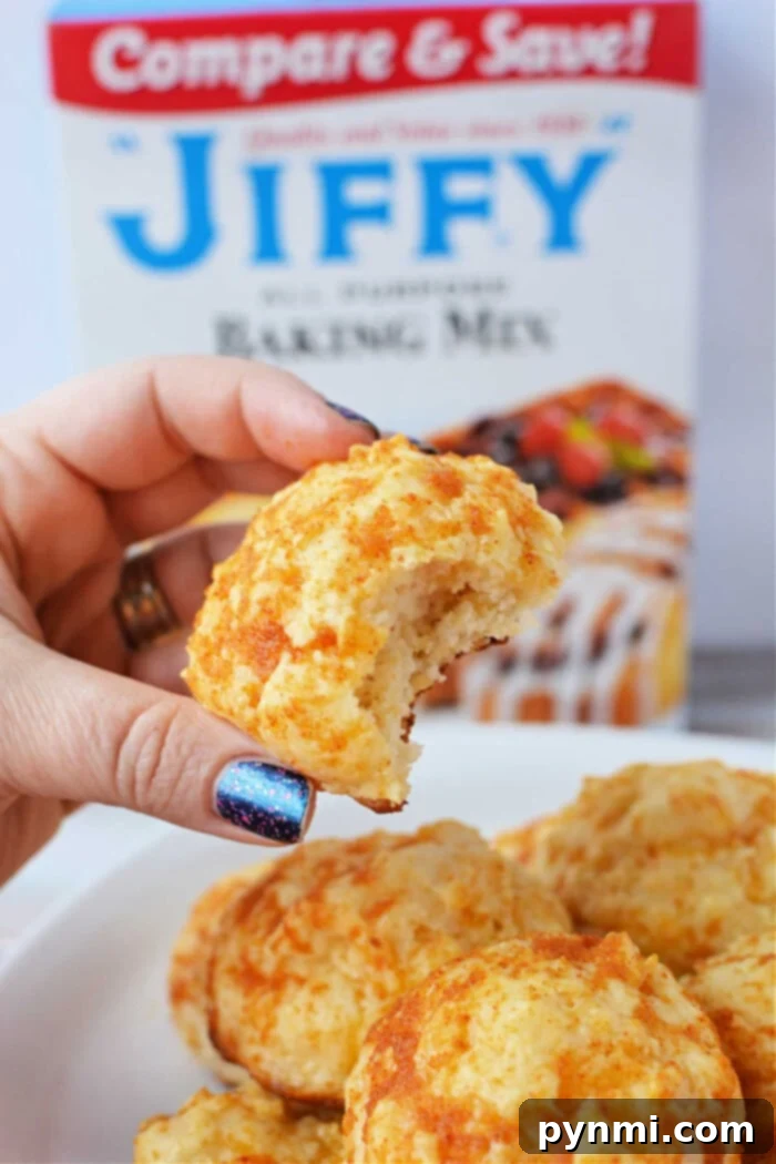 Easy Cheesy Jiffy Biscuits 2 A hand holds a bitten cheddar biscuit, with a Jiffy Mix box adding a tantalizing promise of more flaky treats in the background.