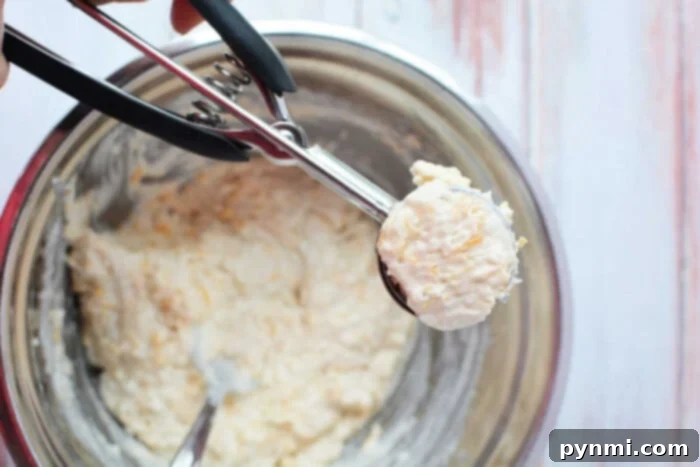 Easy Cheesy Jiffy Biscuits 5 A cookie dough scoop filled with Jiffy Mix dough is held over a mixing bowl on a wooden surface, ready to bake delicious biscuits.