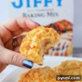 A hand holds a bitten cheddar biscuit, with a Jiffy Mix box adding a tantalizing promise of more flaky treats in the background.