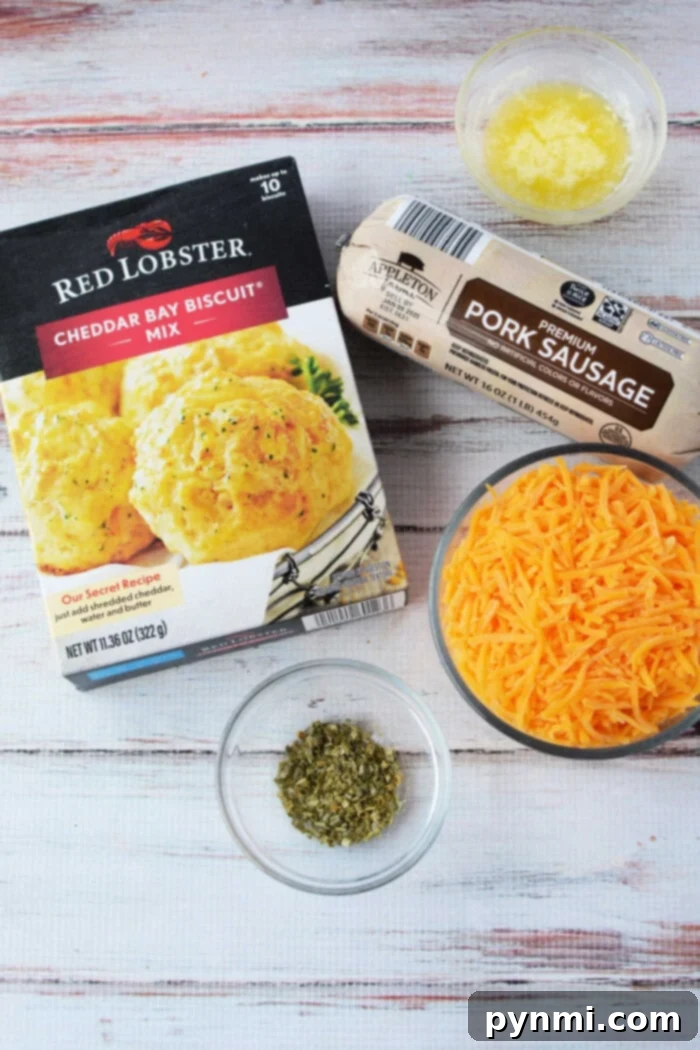 A rustic presentation of key ingredients for sausage balls, including a box of Red Lobster Cheddar Bay Biscuit Mix, a roll of savory pork sausage, and a bowl of finely shredded cheese, ready for preparation.