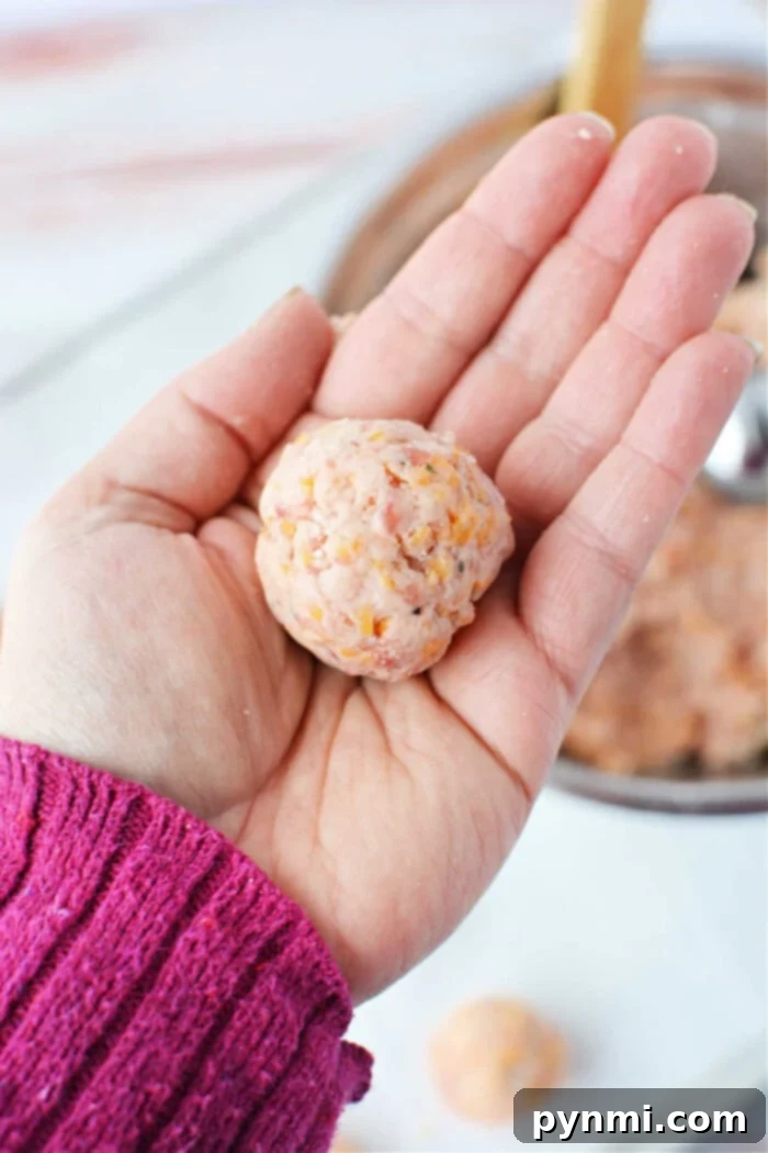 A close-up shot of a hand, adorned with a pink sweater, meticulously rolling a raw sausage ball mixture. The rustic wooden table below holds a bowl brimming with more of the savory ingredients, hinting at the preparation process.