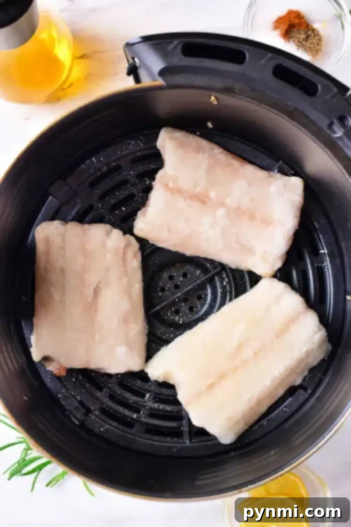 Three frozen cod fillets are placed in an air fryer basket. Nearby, there is a small bowl of spices, a glass bottle of oil, rosemary sprigs, and lemon slices on a light-colored surface.