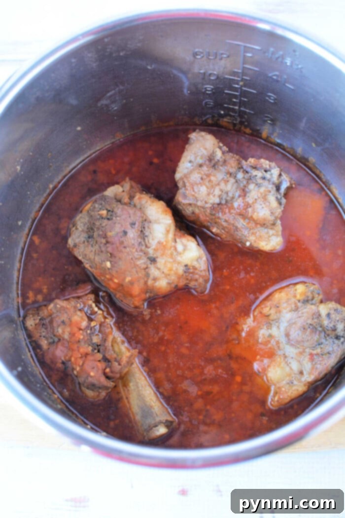 Cooked oxtail in a pot with rich, red broth, hinting at the tenderness achieved by using a pressure cooker. Four pieces are visible alongside aromatic spices and herbs.