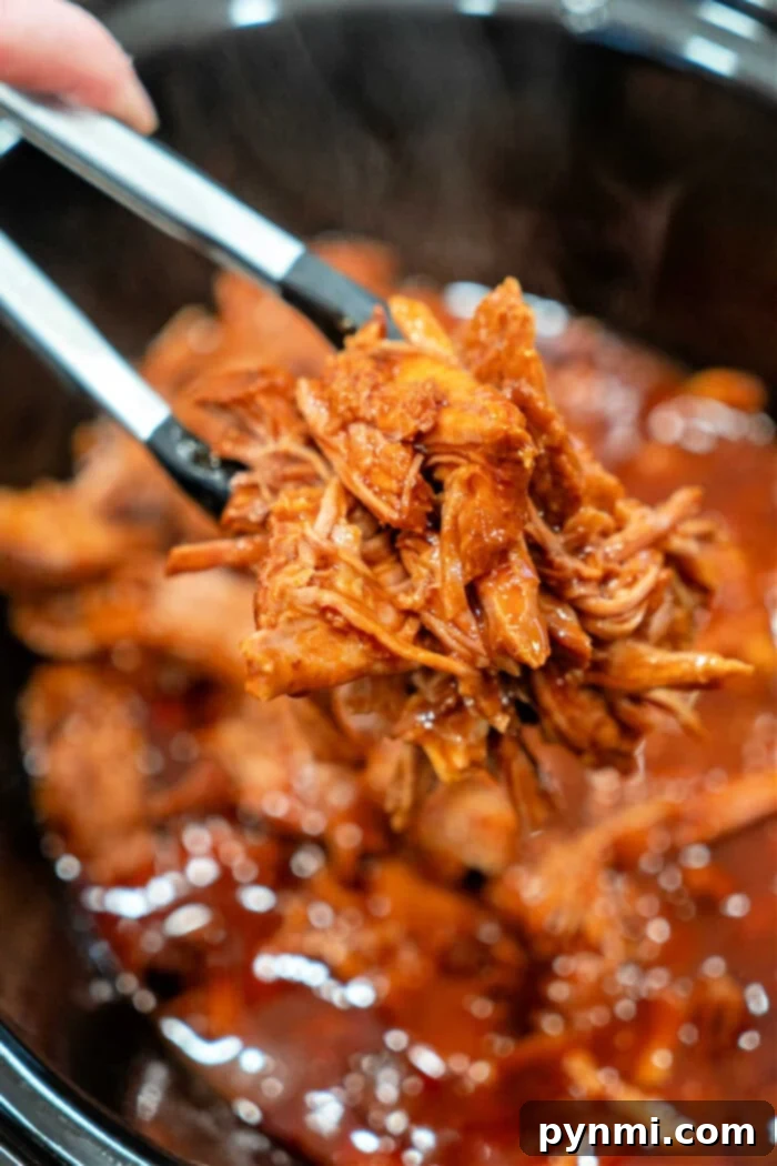 Two-Ingredient Pulled Pork Perfection 2 Tongs holding shredded BBQ pulled pork over a slow cooker, showcasing the perfectly cooked, easy 2-ingredient pulled pork ready to serve.