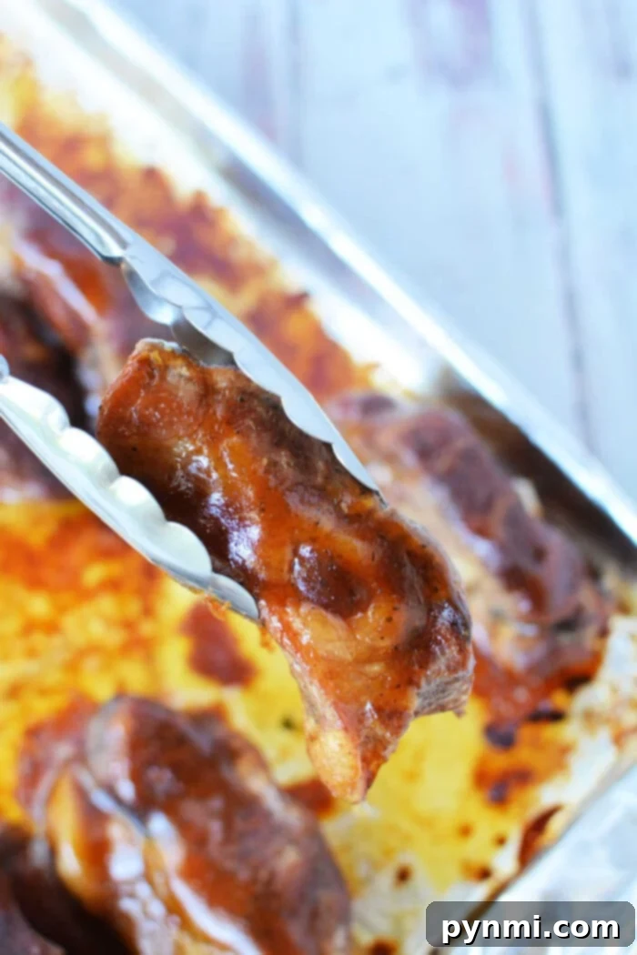 Tongs holding a piece of beautifully glazed boneless pork ribs above a baking tray, ready for the oven, with more succulent, glazed meat waiting beneath.