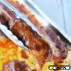 Tongs holding a piece of beautifully glazed boneless pork ribs above a baking tray, ready for the oven, with more succulent, glazed meat waiting beneath.