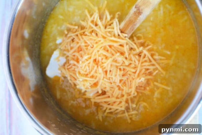 Rich, shredded cheese is stirred into a gently bubbling, creamy yellow potato soup in a pot using a wooden spoon. This hearty Pressure Cooker Potato Soup is made effortlessly with frozen hash browns, providing quick comfort.