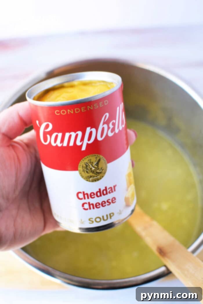 A hand holds a can of Campbell's Cheddar Cheese soup, poised above a deep pot filled with a creamy yellow potato soup, alongside a wooden spoon. This key ingredient is perfect for enhancing the flavor of potato soup made with frozen hash browns in a pressure cooker.