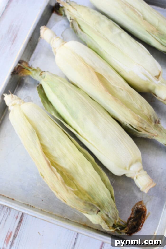 Five unpeeled ears of corn on the cob rest side by side on a metal baking tray, ready for oven cooking, arranged neatly on a wooden surface.