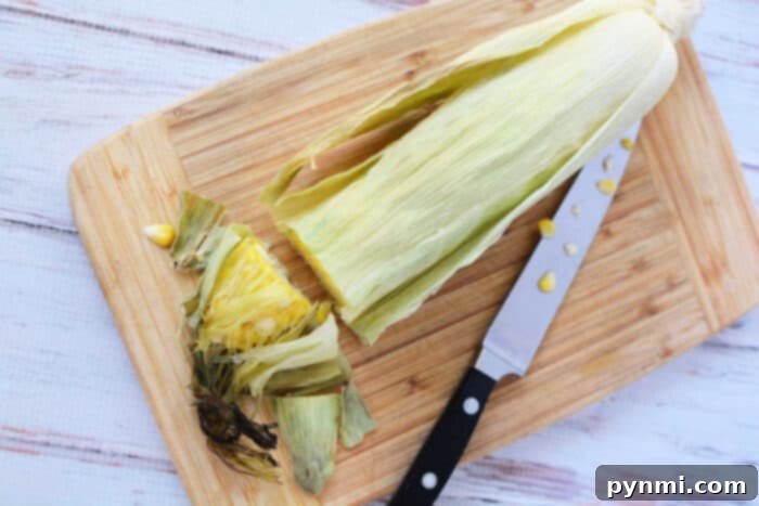 A partially peeled corn cob with husk rests alongside a knife on a wooden cutting board, ready for an adventure in oven cooking.