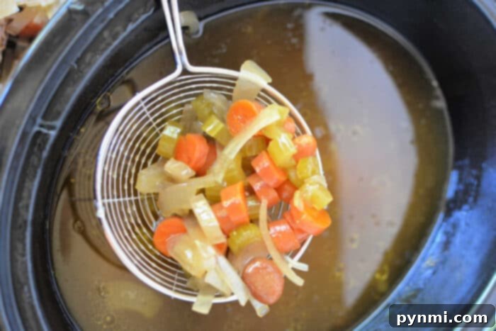 Set It and Forget It Slow Cooker Bone Broth 5 A wire ladle lifts diced carrots, celery, and onions above a pot of simmering bone broth, creating a fragrant medley perfect for your slow cooker.