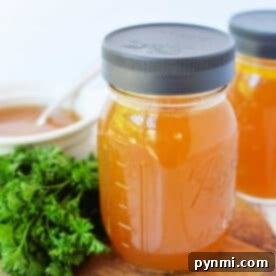 Two mason jars of golden bone broth sit on a wooden board, accompanied by fresh parsley, with a white bowl in the background. Perfect for your favorite slow cooker broth recipe.