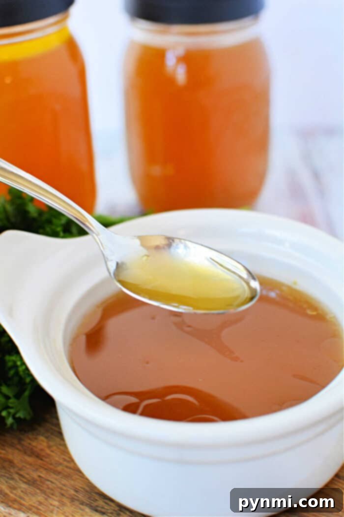 Set It and Forget It Slow Cooker Bone Broth 7 A spoon hovers over a white bowl, filled with homemade bone broth made in a slow cooker, with two jars in the background.