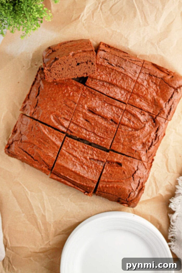 Protein-Packed Perfection Brownies 2 A rich, sliced brownies sits invitingly on parchment paper, a white plate poised nearby, with a hint of greenery in the corner. Try this as a delightful twist on your favorite protein brownie recipe.