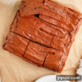 A rich, sliced brownies sits invitingly on parchment paper, a white plate poised nearby, with a hint of greenery in the corner. Try this as a delightful twist on your favorite protein brownie recipe.