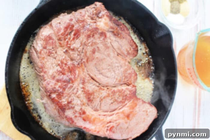 A large boneless chuck roast sizzles in a cast iron skillet, with seasonings and broth beside it.