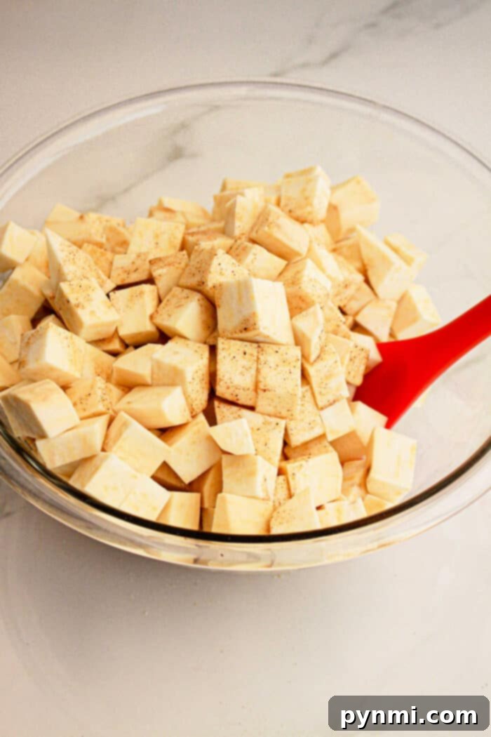 White Sweet Potato Delights 4 Glass bowl of cubed raw sweet potato with seasoning, stirred by a red spatula on a white countertop—perfect for adding to your favorite White Sweet Potato Recipe.