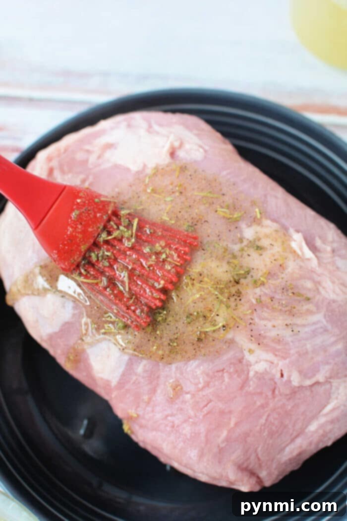 Mouthwatering Slow Cooker Porketta Roast 3 A Crockpot Porketta Roast is prepared as a delectable raw pork roast, with a flavorful marinade gently applied using a red brush on a sleek black plate.