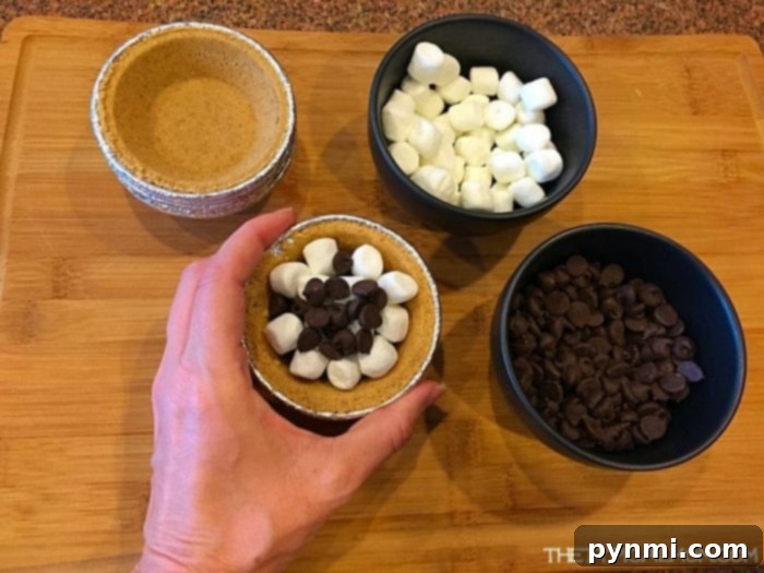 Assembling Mini S'mores Pies for Grilling Perfection A hand carefully placing marshmallows and chocolate chips into a mini graham cracker pie crust, alongside bowls of ingredients, demonstrating an easy S'mores pie recipe for the barbecue.