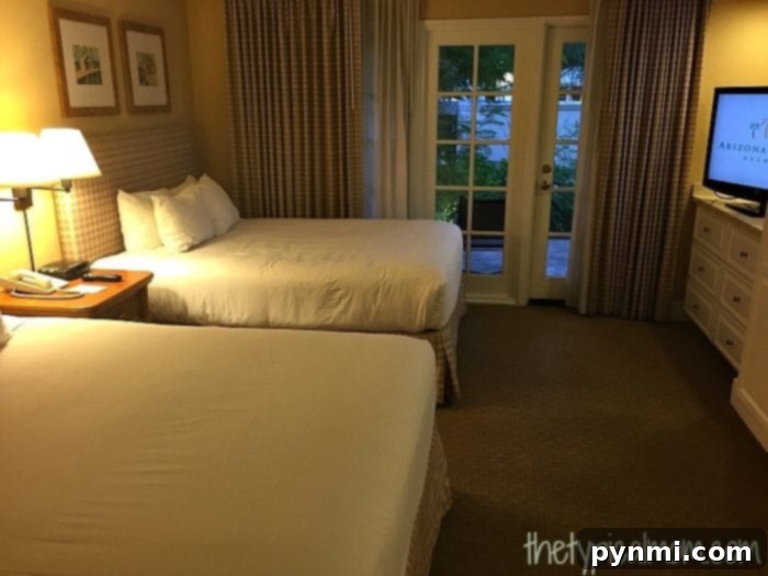 The Arizona Grand Resort offers a hotel room featuring two double beds, a nightstand with a lamp, and a dresser. A glass door with curtains opens to a patio with a serene garden view.