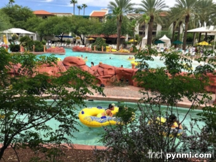 At the Arizona Grand Resort, the outdoor pool area features a lively lazy river where guests relax on inflatable tubes, surrounded by tropical plants and palm trees. Lounge chairs invite visitors to unwind in this sun-soaked paradise.