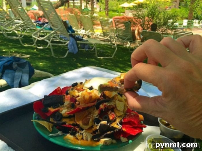 A person at the Arizona Grand Resort grabs a loaded nacho chip from a plate, surrounded by lounge chairs and lush green grass, poolside.