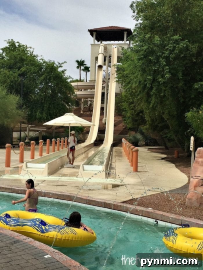 Two people in yellow inflatable rings float along a lazy river at Arizona Grand Resort, nestled at the bottom of two tall water slides. A lifeguard stands watch as trees and umbrellas create a serene oasis around the attraction.