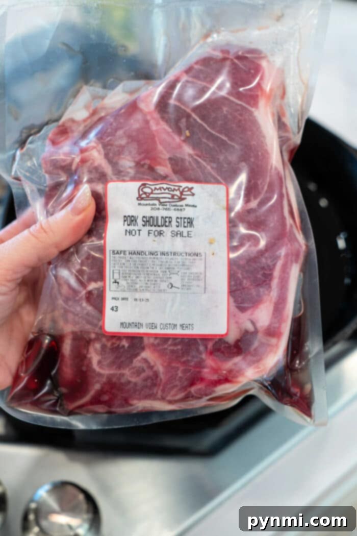 A hand holding a vacuum-sealed package of raw pork shoulder steak, emphasizing its quality and preparation for cooking.