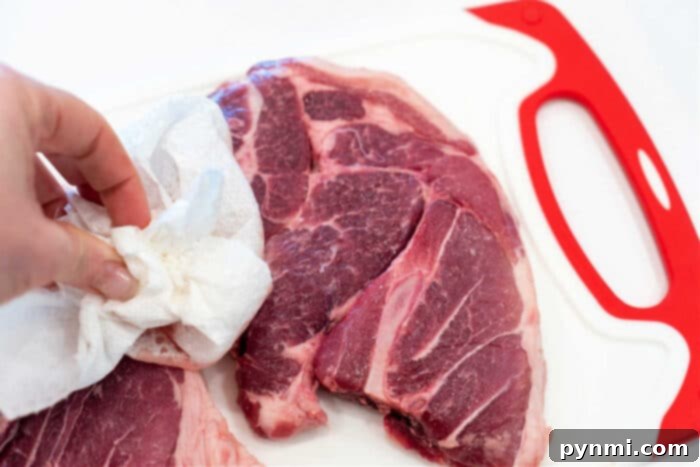 A close-up of a hand dabbing raw pork shoulder steaks with a paper towel on a white cutting board, highlighting the importance of drying the meat for searing.