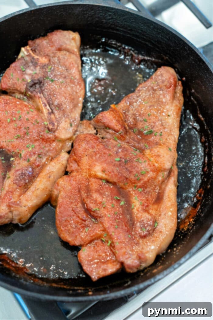 A dynamic shot of two seasoned pork shoulder steaks sizzling and browning in a hot cast iron skillet on a stovetop, capturing the cooking process.