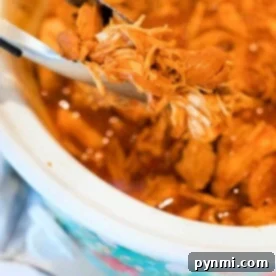 Close-up of pulled chicken being held by tongs above a crockpot. This BBQ chicken recipe showcases tender, shredded meat in a savory sauce, with the slow cooker lined with a floral pattern on the outside.