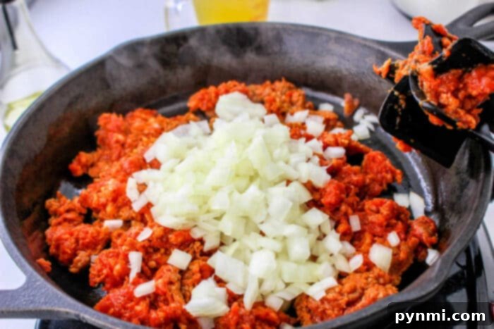 Robust Chorizo Stuffing 3 Chopped onions on top of raw ground meat in a black skillet, being stirred with a spatula—perfect as the base for a flavorful chorizo stuffing or your favorite stuffing recipe.