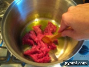A hand uses a wooden spoon to cook ground meat in a pot on the stove, preparing the base for a delicious one pot spaghetti meal.