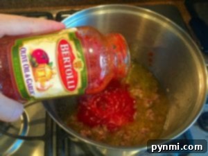 A hand pours Bertolli olive oil and garlic sauce into a pot on the stovetop, setting the stage for a delicious one-pot spaghetti dish.