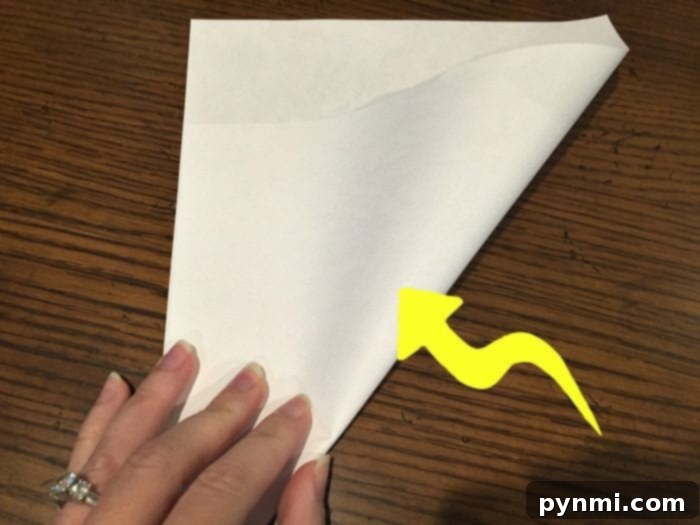 A hand is carefully folding a piece of white paper diagonally on a wooden surface, with a yellow arrow guiding the fold direction, setting the stage for crafting a multiplication fortune teller game.