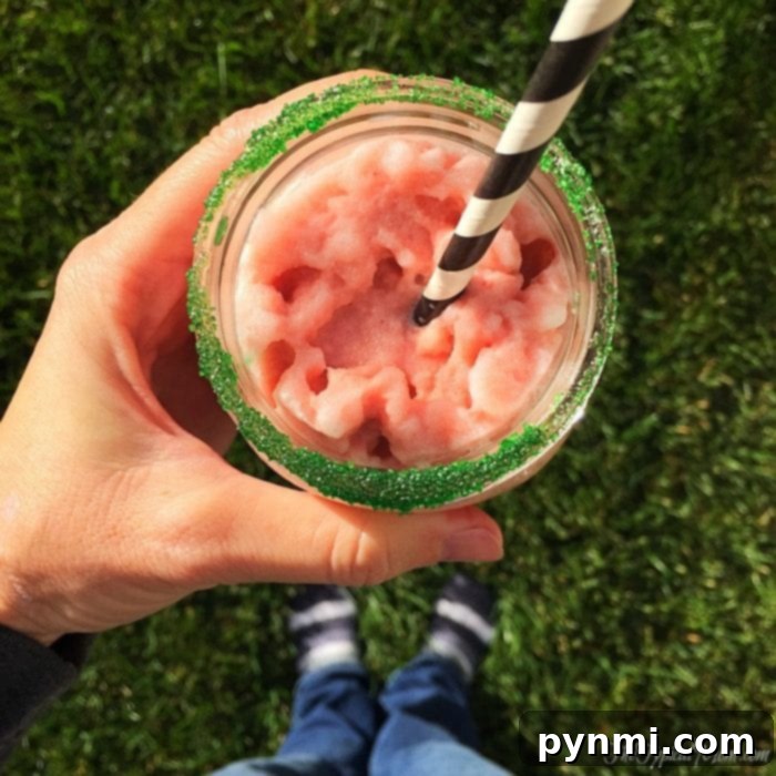 A person enjoys a glass of pink slushy with a black and white striped straw, its rim dusted in green sugar. This homemade peppermint frappuccino offers a festive and refreshing treat.