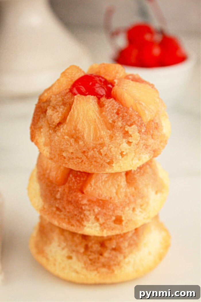 Effortless Golden Pineapple Upside Down Cupcakes 8 Three mini pineapple upside-down cupcakes are stacked on top of each other. Each cupcake features caramelized pineapple slices and a maraschino cherry on top. A small bowl of cherries is blurred in the background.