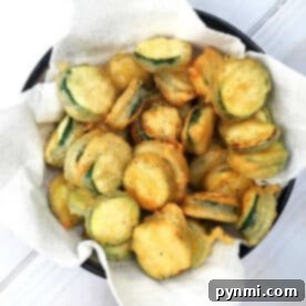 A bowl of crispy salt and pepper zucchini chips rests on a paper towel-lined plate.