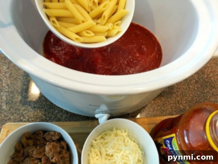 Pasta is added to a slow cooker, mingling with tomato sauce in a symphony of flavors reminiscent of a pizza casserole. Surrounding it are bowls filled with cooked sausage and shredded cheese, eagerly awaiting their turn to join the culinary masterpiece on the counter.