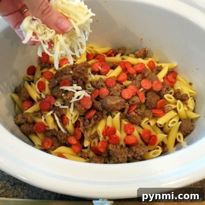 A hand sprinkles cheese over a slow cooker pizza casserole filled with penne pasta, sausage, and pepperoni.